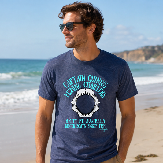 A man wearing a navy blue graphic t-shirt with a shark design. Off shore fishing charters. Fishing shirt on a sandy beach with the ocean in the background.