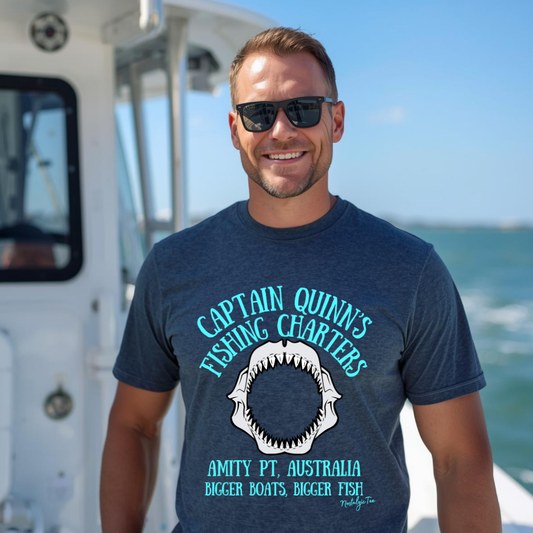 A man wearing a navy blue graphic t-shirt and sunglasses, standing on a boat with the ocean in the background. The t-shirt features a shark mouth graphic and the text 'Captain Quinn's Fishing Charters' and 'Amity Pt, Australia'.
