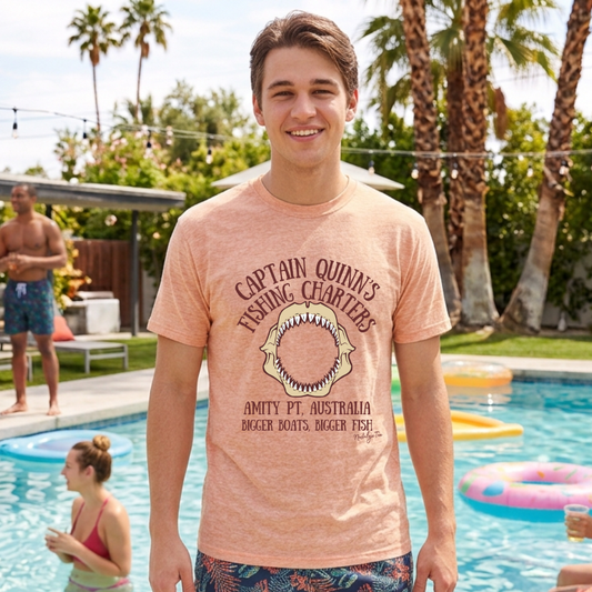 A smiling young man stands in front of a swimming pool wearing a heathered peach t-shirt with a graphic print that reads 'Captain Quinn's Fishing Charters' and 'Amity Pt. Australia'.