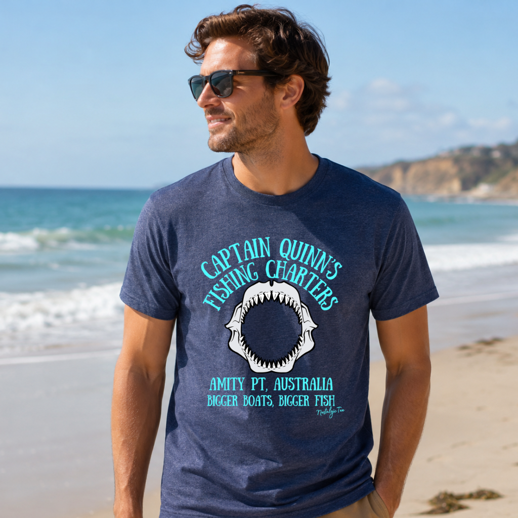 A man wearing a navy blue graphic t-shirt with a shark design. Off shore fishing charters. Fishing shirt on a sandy beach with the ocean in the background.