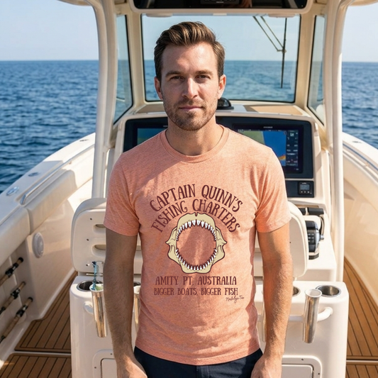 A man wearing a graphic shark tee stands on the helm of a boat with the ocean in the background. The t-shirt features a shark graphic and the text 'Captain Quinn's Fishing Charters'.