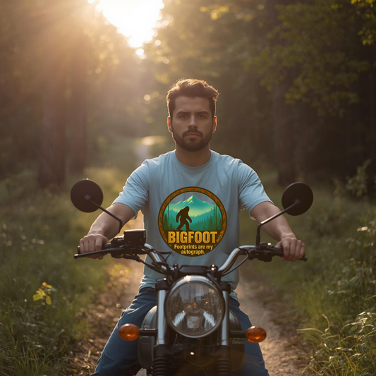 Man riding a motorcycle in a forest wearing a t-shirt with a Bigfoot design.